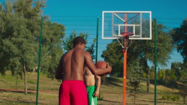 Active fit african streetball player performing step back shot on outdoor basketball court in morning. Positive diverse male friends exercising at urban court while playing half court basketball game.