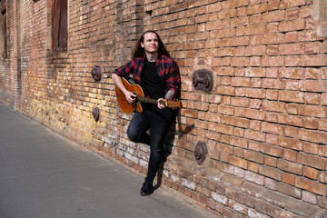 Fototapeta na wymiar Young male with long hair play on acoustic guitar near brown wall 