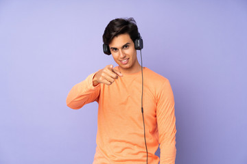 Man over isolated purple background listening music and pointing to the front