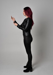 Full length portrait of  girl with red hair wearing black leather jacket, pants and boots. Standing pose  in side profile, isolated against a grey studio background.