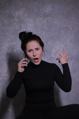 emotional brunette girl in a black turtleneck indignantly talking on the phone on a background of gray wall