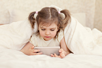 Little girl 3 years old lies under the covers in bed cheerful with smartphone in her hands on white...