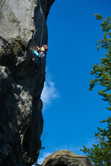 Female rock-climber, climbing up cliff, wearing blue leggings and black top in safety harness, clear blue sky on background. Low angle view. Concept of mountaineering, alpine climbing, extreme sports.