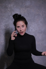 emotional brunette girl in a black turtleneck indignantly talking on the phone on a background of gray wall