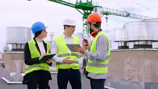 Group of specialists multiethnic on the roof top of construction site using digital tablet to analyzing some details of construction site they wearing safety helmets in front of them standing a big