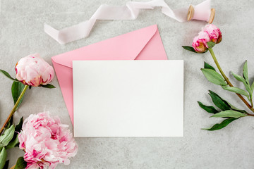 Mockup invitation, blank paper greeting card, pink envelope and peonies on gray stone table. Flower background. Flat lay, top view.