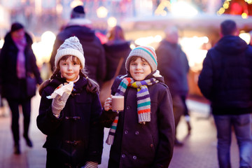 Cute little kids girl and boy having fun on traditional Christmas market during strong snowfall. Happy children eating traditional curry sausage called wurst and drinking hot chocolate. Twins friends