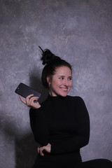 emotional portrait of a happy brunette girl in a black turtleneck holding black phone against a gray wall. High quality photo