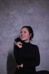 Fototapeta premium emotional portrait of a happy brunette girl in a black turtleneck holding black phone against a gray wall. High quality photo
