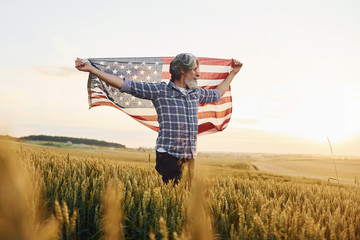 Holding USA flag in hands. Patriotic senior stylish man with grey hair and beard on the agricultural field