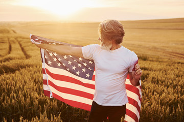 Holding USA flag in hands. Patriotic senior stylish man with grey hair and beard on the agricultural field