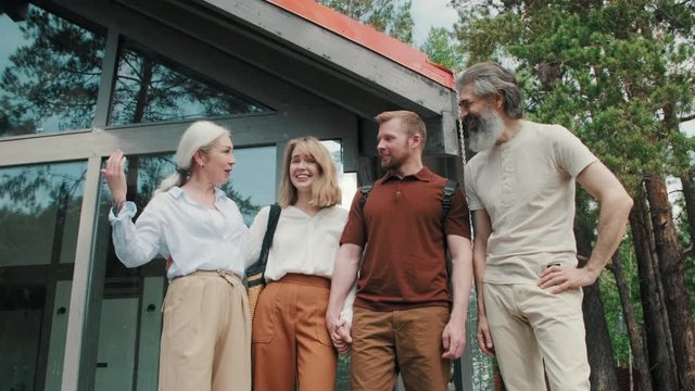 Low Angle Shot Of Happy Elderly Parents And Young Couple Standing Before Modern House With Panoramic Windows And Laughing While Chatting