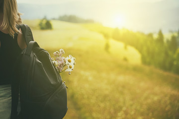 Girl tourist with a backpack and a bouquet of flowers on the background of a mountainous green landscape. View from the back. The atmosphere of summer tourism in nature.Vintage style.