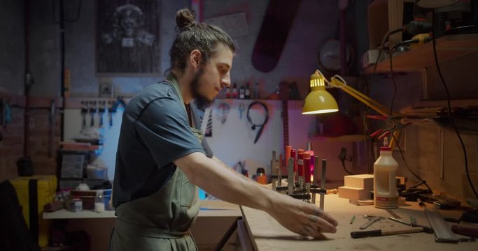 Side shot of handsome Hispanic guy happy enjoying making guitar at cozy workshop indoors, young artisan luther clamps new instrument art craftsman concept hobby of male in apron working in studio