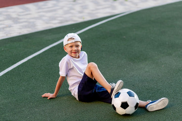 a blond boy in a cap in a sports uniform sits on a football field with a soccer ball, sports section. Training of children, children's leisure