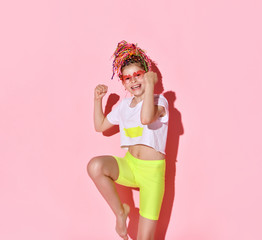 Barefoot girl in neon yellow shorts and white top with rainbow colored african braids raising hands standing on one leg feeling excited