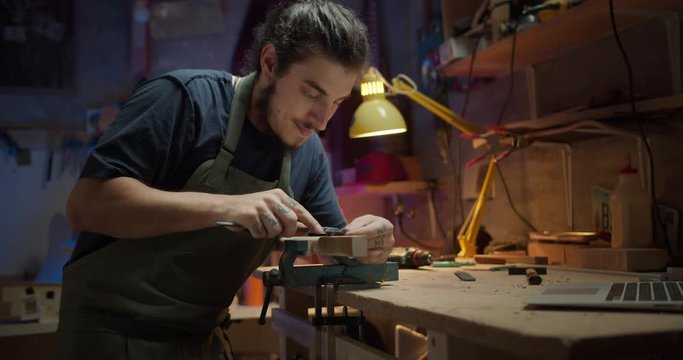 Pleasant artisan male using computer online while making guitar arm in evening workshop, young attractive man in apron using tool instrument working with wooden planks on workbench with laptop