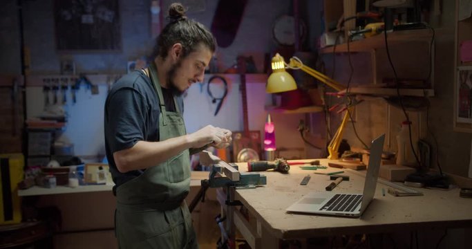 Young handsome luthier beginner searching online using computer how to use instrument, Hispanic male joiner working with wooden planks creating musical guitar instrument at studio in evening