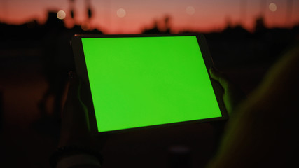 Close up young hands woman uses tablet computer with a horizontal green screen on night street amazing sunset internet map hold pc scroll smart phone social network tourist tracking blank chroma city