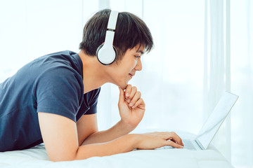 Happy Asian man using Laptop computer and checking good News, Trading, Watching Movie in the Bedroom. COPY SPACE. WORK FROM HOME Concept.