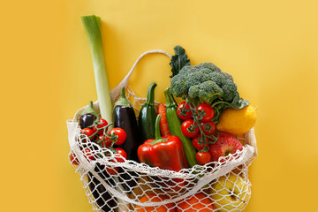 Bunch of mixed organic fruit, vegetables & greens in reusable cotton string net bag. Zero waste concept. Vegan's grocery bag Yellow background, copy space, close up, isolated.