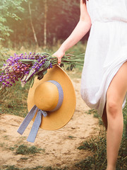 Girl on a village road in a white dress with wild flowers and a summer hat barefoot