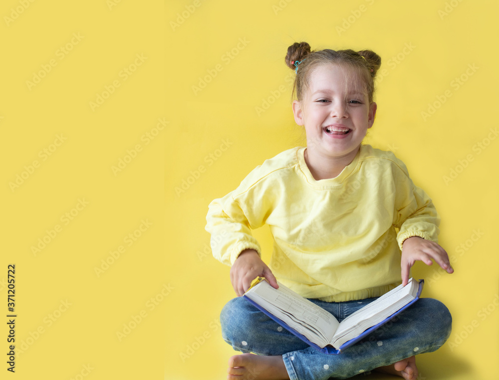 Wall mural funny child girl yellow jacket on a yellow background, sitting, reading a book, development and scho