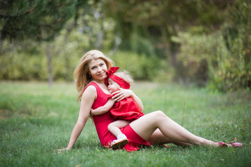 mother and small dauther with blond hair and red dress  sit on green grass smiling