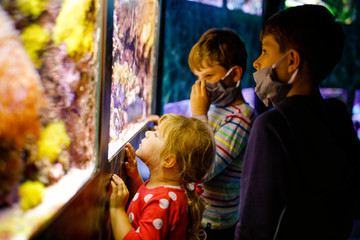 Two kids boys and toddler girl visiting together zoo aquarium. Three children watching fishes and jellyfishes. School boys wearing medicals masks due pandemic corona virus time. Family on staycation - obrazy, fototapety, plakaty