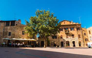 Fototapeta premium Gorgeous view of the Piazza delle Erbe next to the Collegiate Church of Santa Maria Assunta in San Gimignano. A lovely tree stands on the sloping square in the middle between a restaurant and a shop.