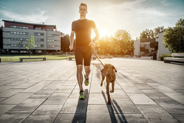 Silhouettes of runner and dog on city street under sunrise sky in morning time. Outdoor walking. Athletic young man with his dog are running in town.