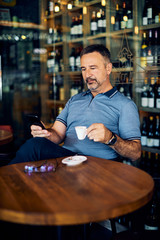 Mature man sitting in coffee shop, drinking coffee and using smart phone.