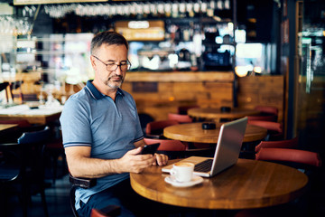 Mature man sitting in coffee shop and using smart phone. There is a coffee and laptop on desk.