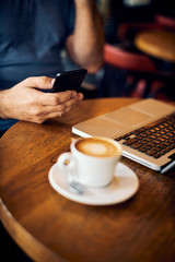 Mature man sitting in coffee shop and using smart phone. There is a coffee and laptop on desk.
