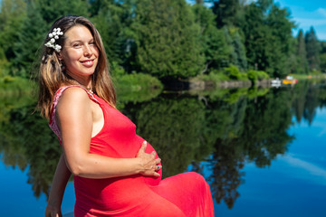 Portrait of a pregnant woman in a sleeveless red dress sitting on the parapet of a lake above the water, caressing her belly and smiling at camera.