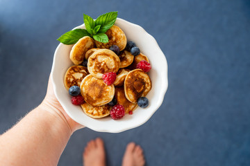 Hand hält Schüssel mit mini Pancakes und frische Beeren. Draufsicht, Barfuss, blauer Fußboden, servierfertig.