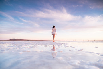 Photo of a slender woman from the back with reflection in the water. A girl walks on a salt lake at sunset.