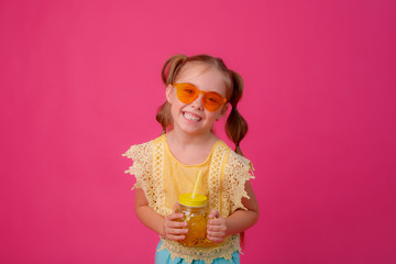 portrait of a little blonde girl in sunglasses on pink with a glass jar of cooling cocktail.