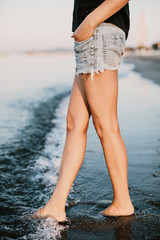 Slim legs of a young and beautiful girl in shorts sit on the sand in the waves of the sea. The waves of the ocean hit the girl's feet. Young ledy is alone on the beach