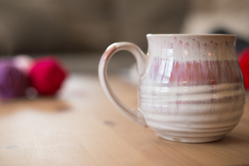 Lightly colored coffee mug with large handle on a pine coffee table with gray sectional sofa in the background, and colorful yarn balls in the foreground.  Cozy day at home with hobbies, no people.