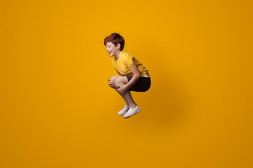 Fototapeta na wymiar Small caucasian boy with red hair smiling and jumping on a yellow studio wall