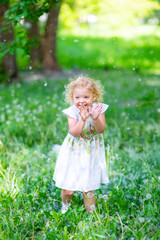 A little girl with white hair and a white dress in the summer in the park stands in a meadow of dandelions