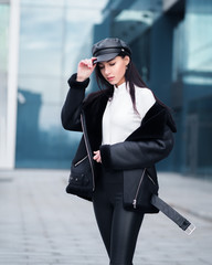 Young brunette woman in the leather pants sheepskin coat and black cappi posing at the city