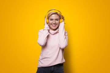 Young blonde woman smiling at camera while listening to music using headphones on a yellow studio wall