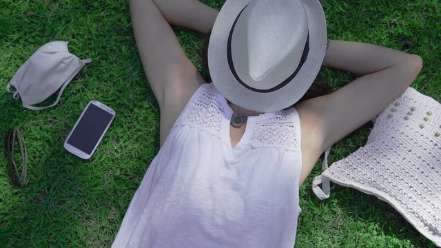 Rotating top view of a Caucasian woman lying on a green field of the park with the face covered by her white hat her white Panama hat and COVID protection mask next to it - City vacation for safety