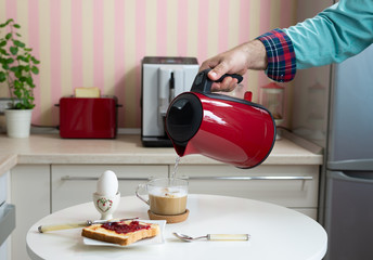 Breakfast served on a kitchen table - boiled egg and jam on bread and with a hand poring hot water from a kettle in a cup of coffee