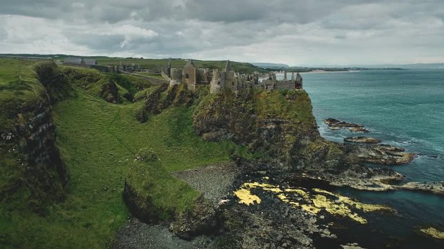Irish castle cliff shore aerial view: Dunluce on green grass valleys and meadows overlooking ocean bay. Beautiful stones and reefs. Beauty of Northern Ireland's historic ruins. Footage shot in 4K, UHD