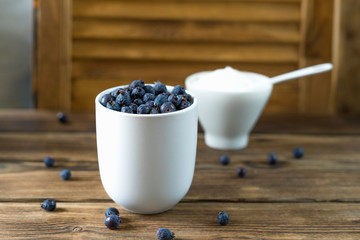 Close up view of a white cup full of fresh shadberry. A cup full of sugar with tea spoon on old grunge wooden table with wooden background. A few berries are scattered on the table. Selective focus