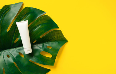 Tube with sunscreen cream on monstera leaf on vivid yellow background, top view. Summer skincare...