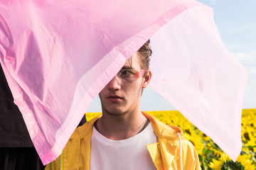 young men wearing a yellow raincoat stands under pink plastic against the backdrop of a field of sunflowers. bright colors, art photo